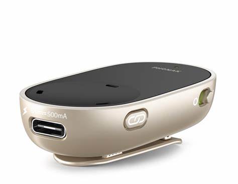 A compact, beige and black assistive listening Phonak PartnerMic device featuring a clip on the bottom for attaching to clothing. The front side has a USB port labeled '500mA' for charging, and a small button marked with a Bluetooth symbol. The device also has an on/off switch on the side and a sleek, curved design.