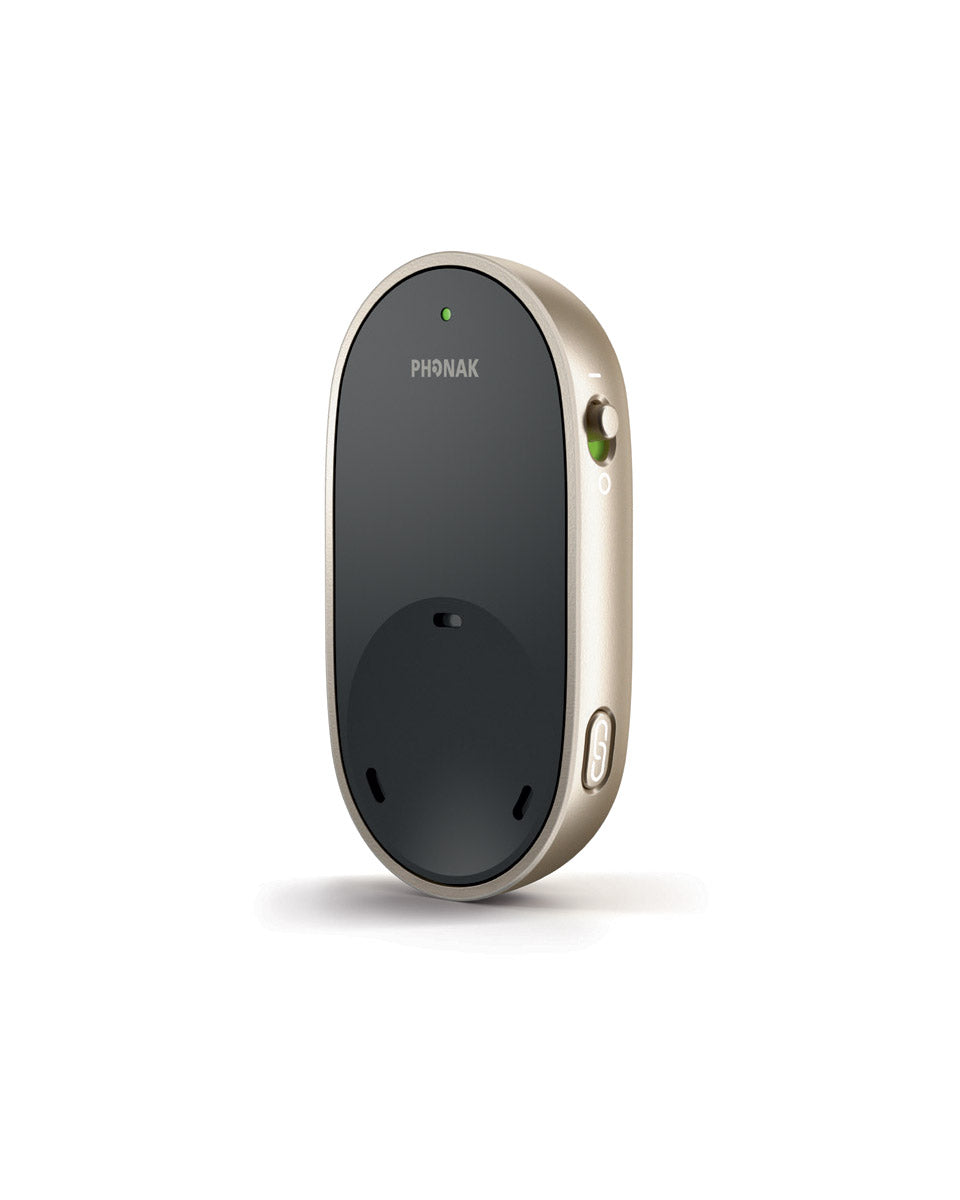 The image depicts a Phonak PartnerMic, a wireless microphone designed to assist people with hearing aids by improving speech understanding in challenging listening environments. The device is oval-shaped, with a sleek black front and gold-colored sides. It has a small green light indicator at the top for power status, a switch on the side to turn it on or off, and a button at the bottom for pairing the microphone to Phonak Hearing Aids.