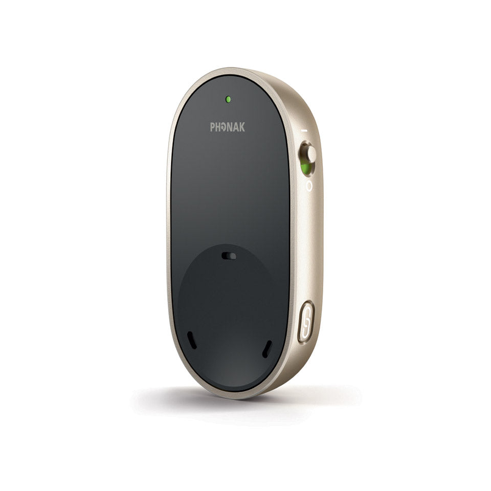 The image depicts a Phonak PartnerMic, a wireless microphone designed to assist people with hearing aids by improving speech understanding in challenging listening environments. The device is oval-shaped, with a sleek black front and gold-colored sides. It has a small green light indicator at the top for power status, a switch on the side to turn it on or off, and a button at the bottom for pairing the microphone to Phonak Hearing Aids.